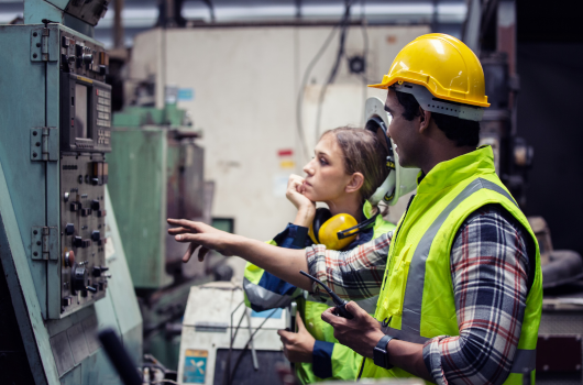 two workers are standing in front of a machine in yellow vests. A male worker is speaking gesturing his hand towards the machine while a female worker listens attentively