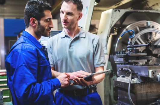 a supervisor holding a clipboard talks to a machinist on the shop floor