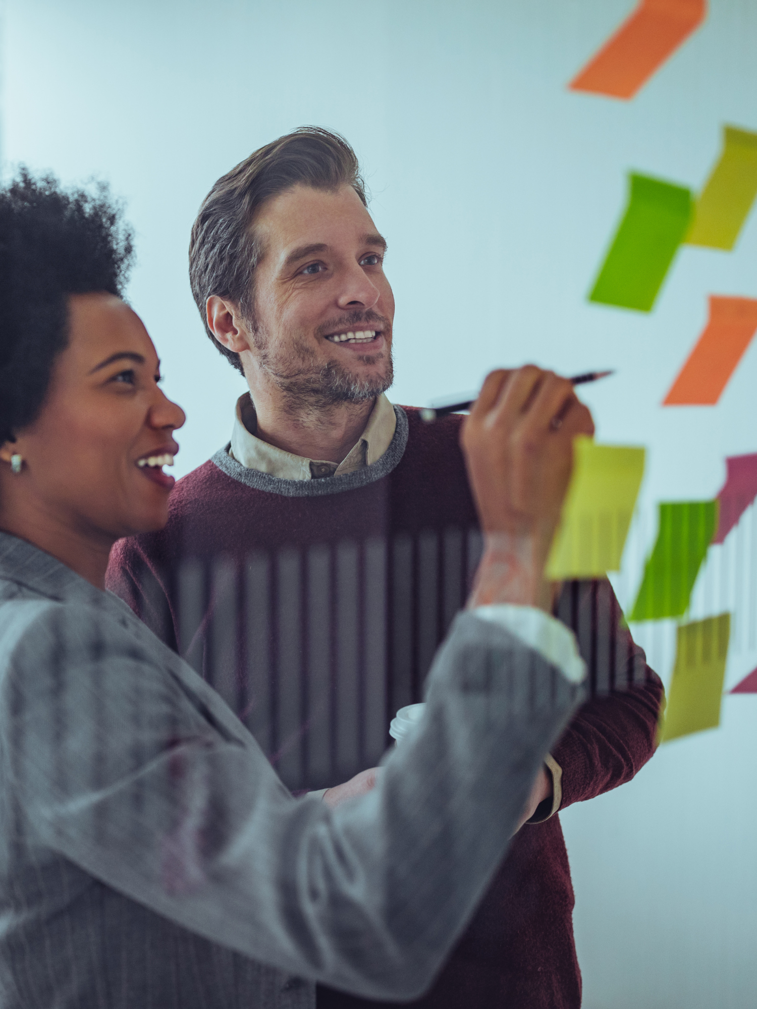 A smiling female holding a pen wearing a gray suit stands next to a smiling male colleague in front of a transparent wall of post-its